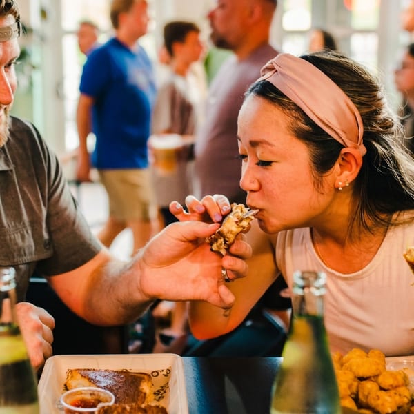 Man feeding a woman a chicken wing at a lively restaurant with other diners in the background.